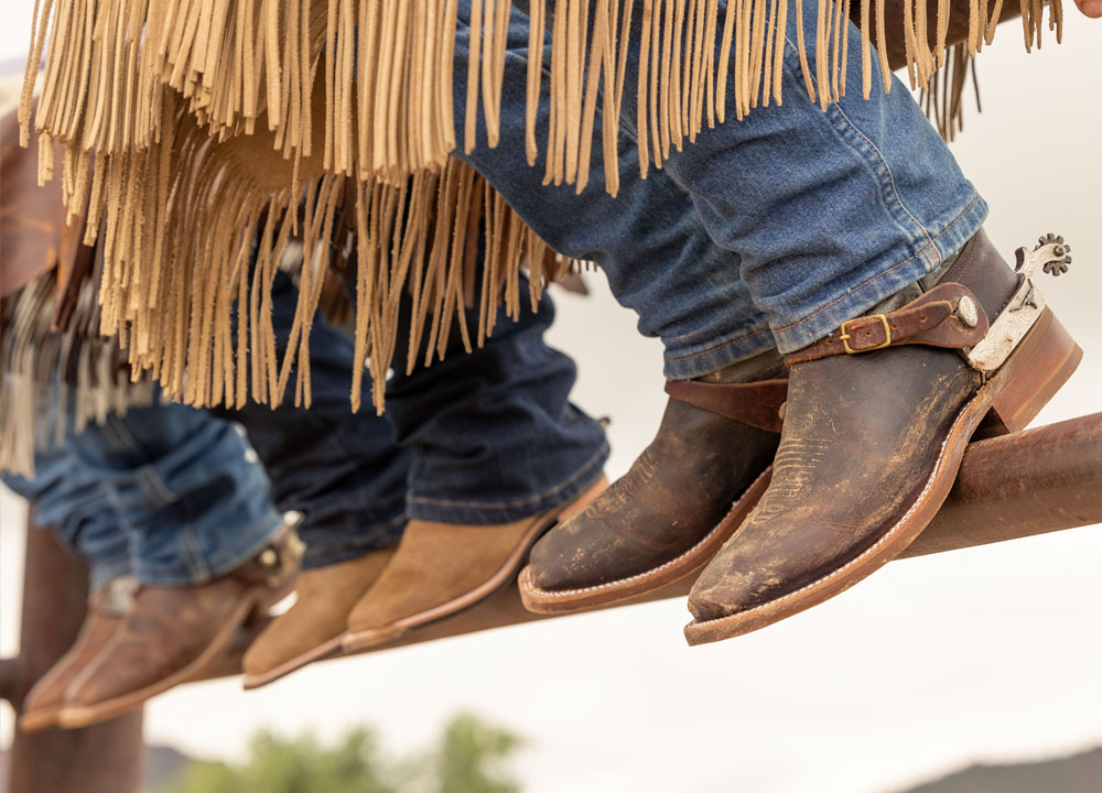 Close up photo of three people sitting on a gate wearing cowboy boots.